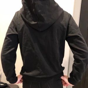 Svart hoodie med dragkedja - Säljer en klassisk svart hoodie med dragkedja och fickor framtill. Tröjan har långa ärmar och en justerbar huva med snören. Perfekt för en avslappnad stil.