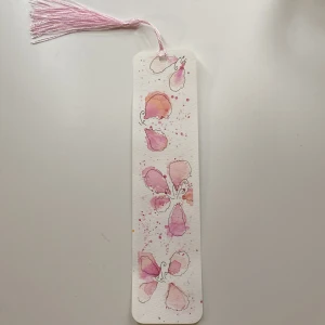 Handmålad bokmärke med blommotiv - Unikt handmålat bokmärke med vackra rosa och orange blommor i akvarellstil. Bokmärket har en rosa tofs upptill och är perfekt för att hålla koll på din plats i boken. En fin detalj för bokälskare.