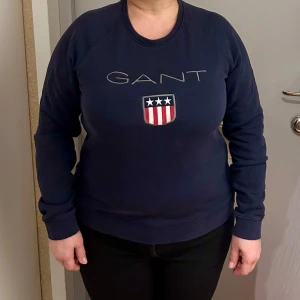 Mörkblå sweatshirt från GANT - Säljer en mörkblå sweatshirt från GANT med deras ikoniska logga och ett emblem på bröstet. Tröjan har långa ärmar och en rund halsringning. Perfekt för en avslappnad stil.