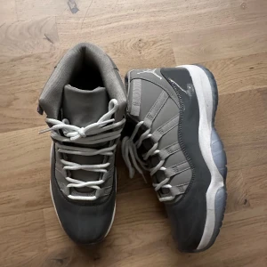 Nike Air Jordans 11 cool grey - Säljer ett par grå Nike Air Jordans med vit sula och snörning. Skorna har en stilren design med hög ankel och tydliga logotyper på baksidan. Perfekta för sneaker-entusiaster!