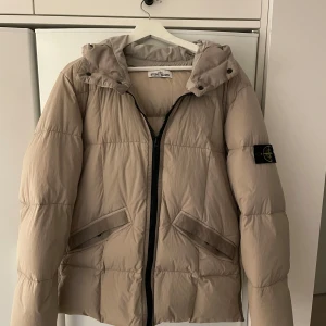Beige dunjacka från Stone Island - Säljer en snygg beige dunjacka från Stone Island.  Den är perfekt för kyligare dagar och har en stilren design och en patch på ärmen. Strl L men ganska liten i storleken.