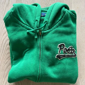 Grön hoodie från Polo Ralph Lauren - Säljer en grön hoodie från Polo Ralph Lauren med dragkedja och broderad logga på bröstet. Den har en klassisk design med huva och långa ärmar. Perfekt för en avslappnad stil.