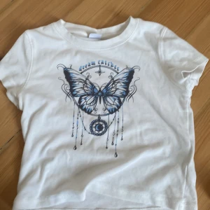 Vit t-shirt med fjärilstryck - Säljer en vit t-shirt med ett snyggt fjärilstryck i blått och texten 'dream catcher'. T-shirten har korta ärmar och en rund halsringning. Perfekt för en avslappnad stil.
