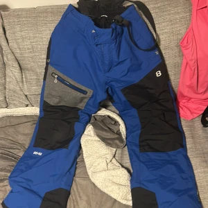 Blå byxor från 8848 Altitude - Säljer ett par blå skidbyxor från 8848 Altitude med svarta och grå detaljer. SkidByxorna har dragkedja och knappar samt justerbara hängslen. Perfekta för utomhusaktiviteter med slitstarkt material och praktiska fickor.