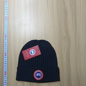 Svart CANADA GOOSE mössa - Snygg svart ribbad mössa med ett runt märke framtill. Perfekt för kalla dagar och ger en stilren look. Mössan är enkel att matcha med olika outfits.
