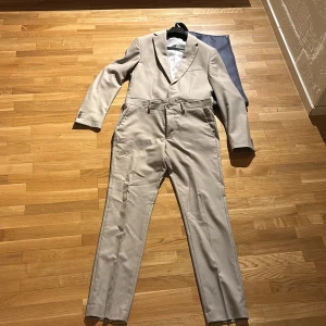 Beige kostym från Riley - Snygg beige kostym från Riley i slim fit. Kavajen har klassiska slag och knappar vid ärmsluten. Byxorna har en stilren design med hällor för bälte och en diskret dragkedja. Perfekt för en elegant look. Perfekt till skolavslutning 😃nypris runt 3000kr, använd 1 gång