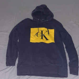 Mörkblå hoodie från Calvin Klein Jeans - Säljer en snygg mörkblå hoodie från Calvin Klein Jeans med en stor gul logga på framsidan. Tröjan har en klassisk passform med långa ärmar och en praktisk magficka. Perfekt till våren. Nypris 1199