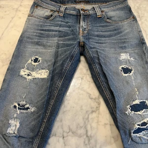 Nudie jeans med slitningar - Säljer ett par blå jeans från Nudie Jeans Co med coola slitningar och en klassisk femficksdesign. Jeansen har en knappgylf och orange sömmar som ger en snygg kontrast. Perfekta för en avslappnad stil.