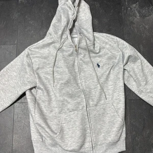 Grå hoodie från Ralph Lauren - Säljer en stilren grå hoodie från Ralph Lauren med dragkedja och justerbar huva. Den har en liten broderad logga på bröstet och är tillverkad i mjukt material för extra komfort. Perfekt för en avslappnad look. O använd 