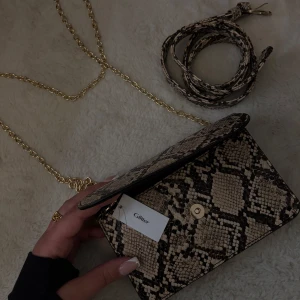 Ormmönstrad axelväska med guldkedja - Snygg axelväska med ormmönster och guldkedja från Glitter. Väskan har en praktisk storlek och ett lock med magnetknäppning. Den medföljande axelremmen är justerbar för bästa passform. Prislapp finns kvar aldrig använd. 