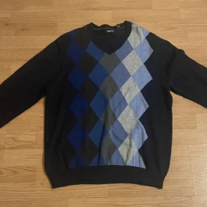 Svart tröja med rutor från Claiborne - Snygg svart tröja från Claiborne med ett stilrent rutmönster i blått och grått framtill. Tröjan har en v-ringad hals och är tillverkad i 100% bomull, vilket gör den både bekväm och lätt att bära. Perfekt för en avslappnad men ändå elegant look.