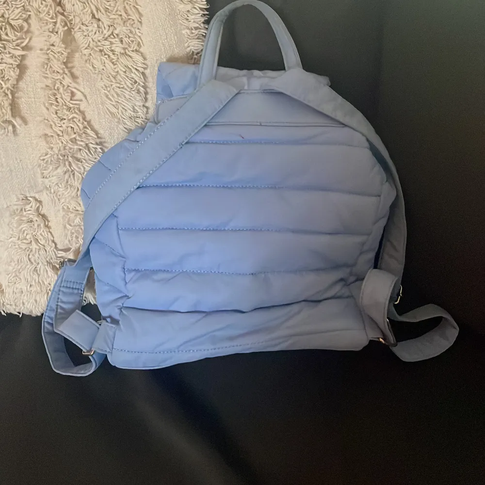 Säljer en snygg blå quiltad ryggsäck med dragkedja och snörning. Perfekt för att bära dina saker med stil. Ryggsäcken har justerbara axelremmar och en praktisk ficka framtill.. Laukut & Käsilaukut.
