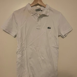Vit pikétröja från Lacoste - Säljer en klassisk vit pikétröja från Lacoste i storlek S. Tröjan är tillverkad i 100% bomull och har korta ärmar samt den ikoniska krokodilloggan på bröstet. Perfekt för en stilren och avslappnad look.