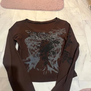 Brun långärmad topp med mönster - Snygg brun långärmad topp med ett unikt mönster i svart och grått. Toppen har en v-ringad design och är dekorerad med små pärlor som ger en extra touch. Perfekt för att ge din outfit en cool stil.