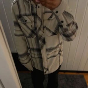 Beige och svart rutig jacka från Bluewear - Säljer en snygg och bekväm beige och svart rutig jacka från Bluewear. Jackan har en huva med dragsko och två bröstfickor med knappar. Insidan är fodrad med mjuk fleece för extra värme.