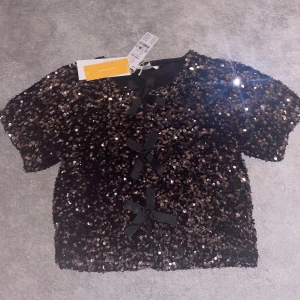 Svart paljettopp med rosetter - Snygg svart paljettopp med korta ärmar och rosetter framtill. Jätte fin och passar till det mesta. Från gina young, aldrig använd. Pris går att diskutera