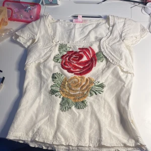 Vit blus med broderade rosor från Moiselle - Charmig vit blus från Moiselle med korta ärmar och vackra broderade rosor i rött och gult på framsidan. Perfekt för att ge en romantisk touch till din outfit. 💐