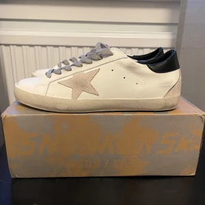Golden Goose Superstar  - Feta sneakers från Golden Goose med en ikonisk stjärndetalj i beige på sidan. Skorna har grå snörning och en svart häl. Perfekta för en stilren och avslappnad look. Endast använd 2 gånger och är i princip i nyskick.