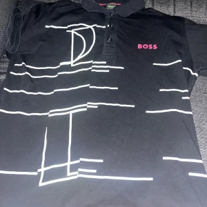 Svart t-shirt från Boss med tryck - Snygg svart t-shirt från Boss med ett unikt vitt tryck och rosa logga på bröstet. T-shirten har en klassisk krage med knappar och är kortärmad. Perfekt för en stilren look. Passar även M