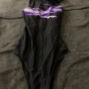 Svart baddräkt från SOC - Snygg svart baddräkt från SOC med ett coolt lila mönster över bröstet. Perfekt för simträning eller en dag på stranden. Baddräkten har en sportig design och ger en bekväm passform.