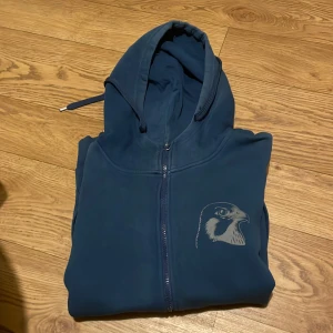 Mörkblå hoodie med Falcon tryck - Säljer en mörkblå hoodie med dragkedja och ett stilrent fågeltryck på bröstet. Tröjan har långa ärmar och en praktisk huva med dragsko. Perfekt för en avslappnad stil.