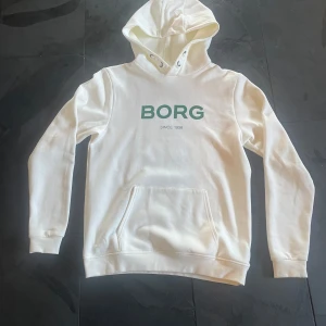Hoodie från Björn Borg - Gräddvit hoodie med grön logga från Björn Borg på framsidan. En rymlig kängurufick på framsida, tvättad endast en gång. En liten fläck som knapps syns som inte går bort.
