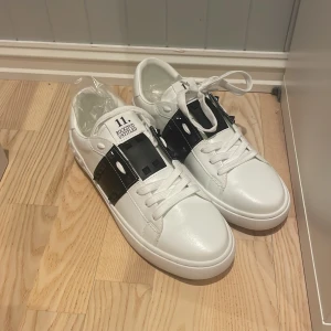 Vita sneakers från Valentino Garavani - Snygga vita sneakers från Valentino Garavani Rockstud 11. med svarta detaljer och snörning. Skorna har en unik design med nitar på hälen och en bred rem över vristen. Perfekta för att ge din outfit en lyxig touch. De är storlek 43 och kommer med påse och dammpåsar.