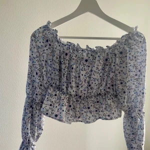 Blommig offshoulder blus från NLY - Säljer en söt blommig offshoulder blus från NLY i vitt med blå och lila blommor. Blusen har långa ärmar och volangdetaljer. Perfekt för en somrig/vårig dag. Mjukt material och smickrande passform! Använd fåtal gånger! 