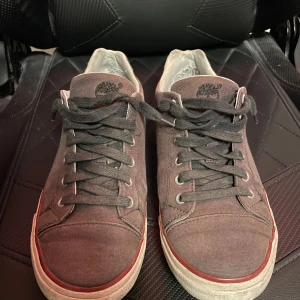 Sneakers från Timberland - Snygga mörkgrå sneakers från Timberland med snörning och vit sula. Skorna har en röd randdetalj runt sulan och är perfekta för en avslappnad stil. Tillverkade i slitstarkt material med Timberland-logga på sidan och sulan. Slitna där bak inuti och på högra sulan där fram, se sista bilden, går at fixa ganska lätt. Annars i fint skick! Hör av er vid funderingar! ( Dessa skor säljs inte längre) 