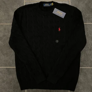 Svart stickad tröja från Polo Ralph Lauren - Snygg svart stickad tröja från Polo Ralph Lauren med det ikoniska röda broderade logotypen på bröstet. Tröjan har en klassisk rund hals och ribbade muddar vid ärmslut och nederkant. Perfekt för en stilren look.