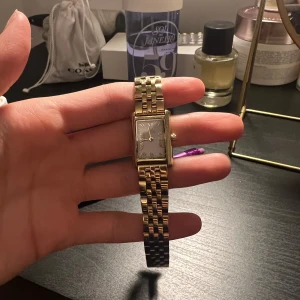 Guldfärgad klocka från Mockberg - Elegant guldfärgad klocka från Mockberg med rektangulär urtavla och romerska siffror. Armbandet är i metall med en klassisk länkdesign. Perfekt för att ge en touch av lyx till din stil.