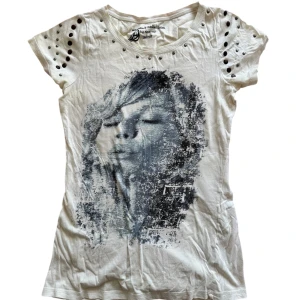 Vit t-shirt med tryck och nitar - Cool vit t-shirt med ett stort svartvitt tryck av ett ansikte på framsidan. T-shirten har korta ärmar och är dekorerad med nitar på axlarna för en edgy look. Perfekt för att ge din outfit en unik touch!