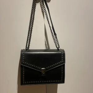 Svart axelväska med nitar och kedja  - Snygg svart axelväska med kedjeaxelrem och coola nitar längs kanterna. Väskan har ett stilrent lås framtill och en praktisk ficka på baksidan. 