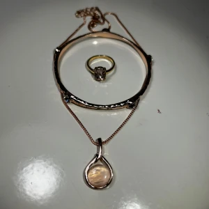 Elegant smyckeset med armband, ring och halsband - Snyggt smyckeset med ett armband, en ring och ett halsband. Armbandet har en unik textur och små stenar. Halsbandet har en kedja med ett droppformat hänge. Perfekt för att ge en elegant touch till din stil.