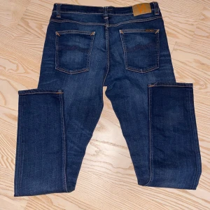Mörkblå jeans från nudie - Snygga mörkblå jeans från nudie med klassisk femficksdesign och orange kontrastsömmar. De har en dragkedja och knapp framtill. Perfekta för en avslappnad stil. Storlek 32/34