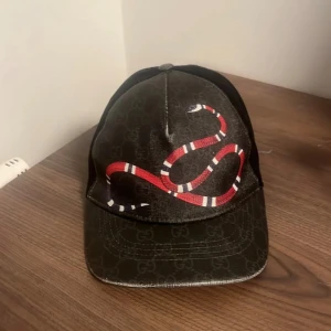 Svart keps med orm från Gucci - Snygg svart keps från Gucci med ett coolt ormmotiv i rött och vitt på framsidan. Kepsen har ett justerbart band med röd och blå rand på baksidan. Perfekt för att ge din outfit en lyxig touch. Den är ett fåtal gånger använd byter endast mot tiger eller orginal med blå och beige