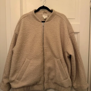 Beige teddyjacka från H&M - Mysig beige teddyjacka från H&M med dragkedja framtill. Jackan har en avslappnad passform och två praktiska fickor. Perfekt för kyliga dagar när du vill hålla dig varm och stilren.