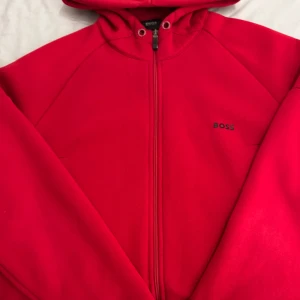Röd huvtröja från Boss - Säljer en snygg röd huvtröja från Boss med dragkedja framtill. Tröjan har en stilren design med en liten logotyp på bröstet och en bekväm huva. Perfekt för en casual look. Väldigt liten i storlek så de kanske är en L egentligen! billig och pris går att diskuteras, 