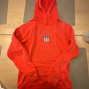 Röd hoodie från GANT - Säljer en snygg röd hoodie från GANT med ett tryckt emblem på bröstet. Storlek 158/164. Mycket bra skick, Kan fraktas eller hämtas i Skövde mvh Lukas. 