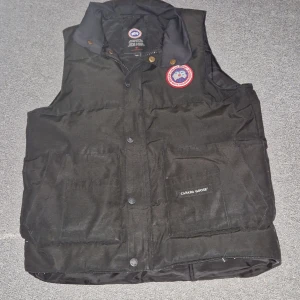 Svart dunväst från Canada Goose - Säljer en svart dunväst från Canada Goose med klassisk design och varumärkets logga på bröstet. Västen har knappar och dragkedja framtill samt praktiska fickor. Perfekt för kyligare dagar.