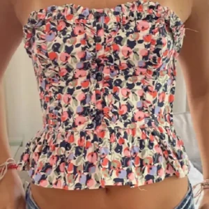Blommig tubtopp med volang - Säljer en söt blommig tubtopp med volangdetaljer. Toppen har ett färgglatt mönster i rosa, blått och vitt. Perfekt för sommardagar!