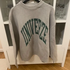 Grå tröja med text - Säljer en grå tröja med texten 'UNIVERZE' i grönt på framsidan. Tröjan har en ribbad krage och långa ärmar, perfekt för en avslappnad stil. Passar bra till jeans eller leggings.