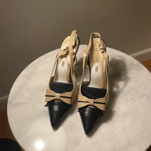 Beige och svarta klackskor från Peter Kaiser - Eleganta klackskor från Peter Kaiser i beige och svart. Skorna har en spetsig tå och en dekorativ rosett framtill. De är tillverkade i skinn och har en smal klack som ger en stilren look.