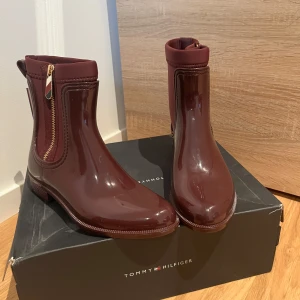  boots från Tommy Hilfiger - Snygga vin röda boots från Tommy Hilfiger i glansigt material. De har en dragkedja på sidan för enkel på- och avtagning. Perfekta för en stilren look.