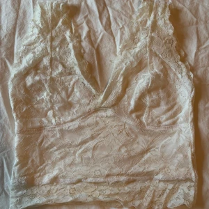 Vit spetslinne - Säljer en vacker vit spetslinne med v-ringad design. Toppen är ärmlös och har ett fint blommigt mönster i spetsen. Perfekt för att lägga till en romantisk touch till din outfit.