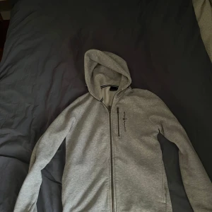 Grå hoodie från Sail Racing - Säljer en stilren grå hoodie från Sail Racing med dragkedja framtill. Den har en klassisk design med huva och långa ärmar. Den är i använt skick så skulle säga typ 7/10! Bara att höra sig vid frågor eller funderingar!
