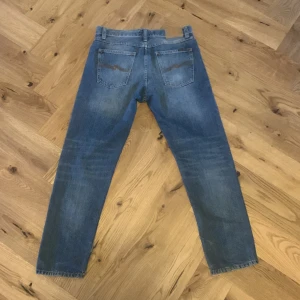 Blå jeans från Nudie Jeans - Snygga blå jeans från Nudie Jeans med klassisk femficksdesign och orange sömmar på bakfickorna. De har en knappgylf och är tillverkade i slitstarkt denim. Perfekta för en avslappnad stil. Modellen är steady eddie