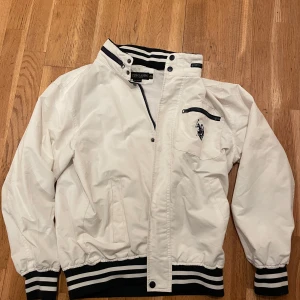 Vit jacka från U.S. Polo Assn. - Säljer en stilren vit jacka från U.S. Polo Assn. med svarta och vita randiga detaljer vid krage, ärmslut och nederkant. Jackan har dragkedja och knappstängning samt en broderad logga på bröstet. Perfekt för en våren/hösten. Den är XL i barn strlk, vilket är S i vuxen strlk. Skick 6/10
