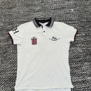 Vit pikétröja från Adrian Hammond - Snygg vit pikétröja från Adrian Hammond med broderade detaljer och tryck. Tröjan har korta ärmar med röd och svart kant, samt en krage med text. Perfekt för en stilren look.