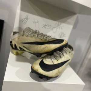 Nike mercurial elit  - Snygga beige fotbollsskor från Nike med svart swoosh och snörning. Skorna har en sportig design med en slitstark sula, perfekt för fotbollsplanen.